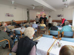 Prebivalci doma starejših občanov sedijo za mizami in poslušajo predavanje.